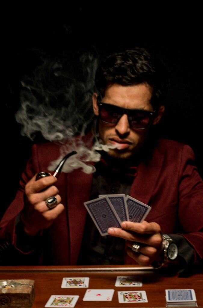 A mysterious man wearing sunglasses plays poker, holding cards and a smoking pipe in dramatic lighting.